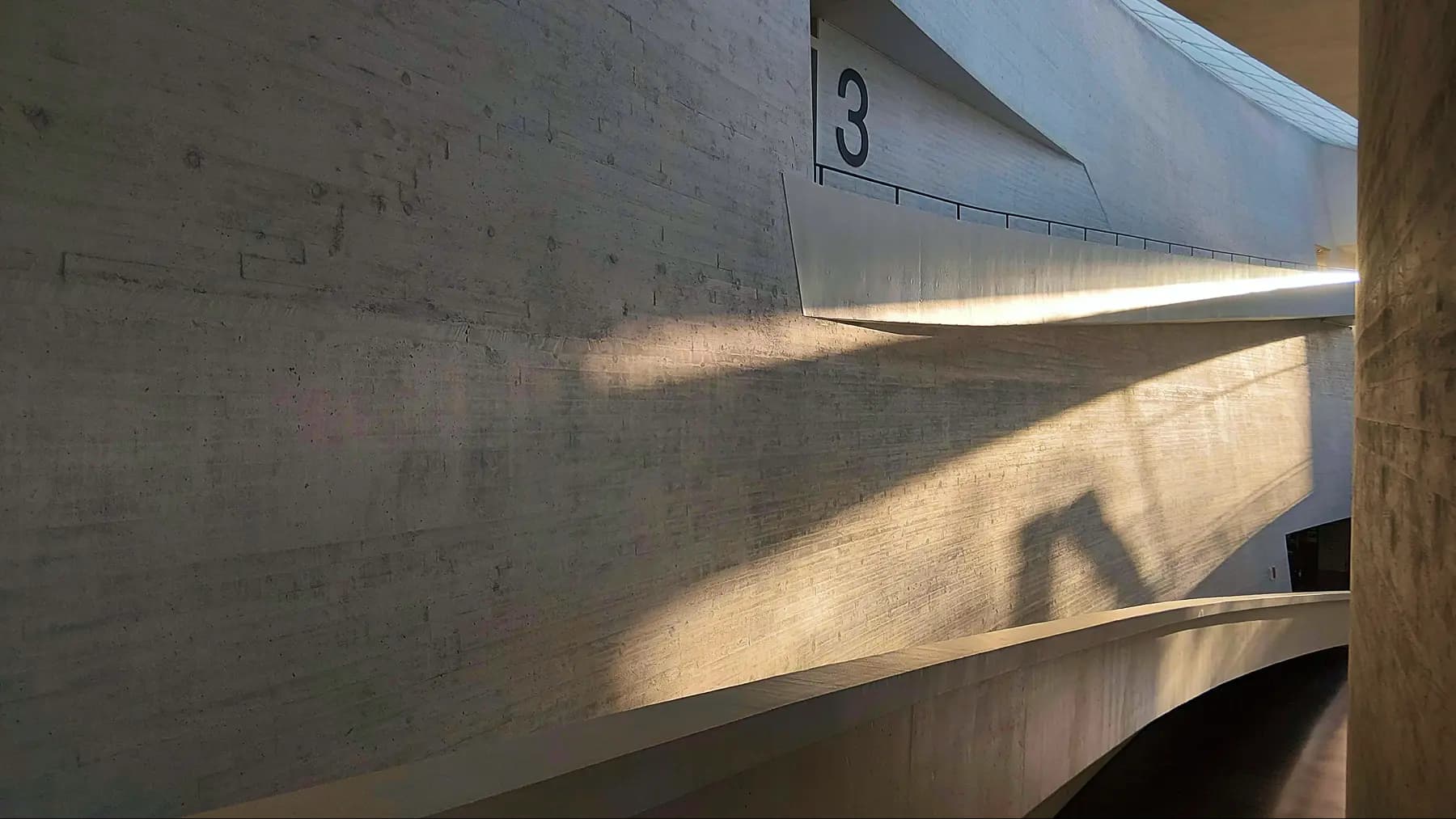 Curved concrete hallway with sunlight casting angular shadows and number 3 on the wall.